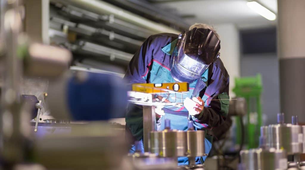 A welder in protective gear works on metal components on an industrial factory floor, illustrating the operational complexity that defines industrial executive search mandates.