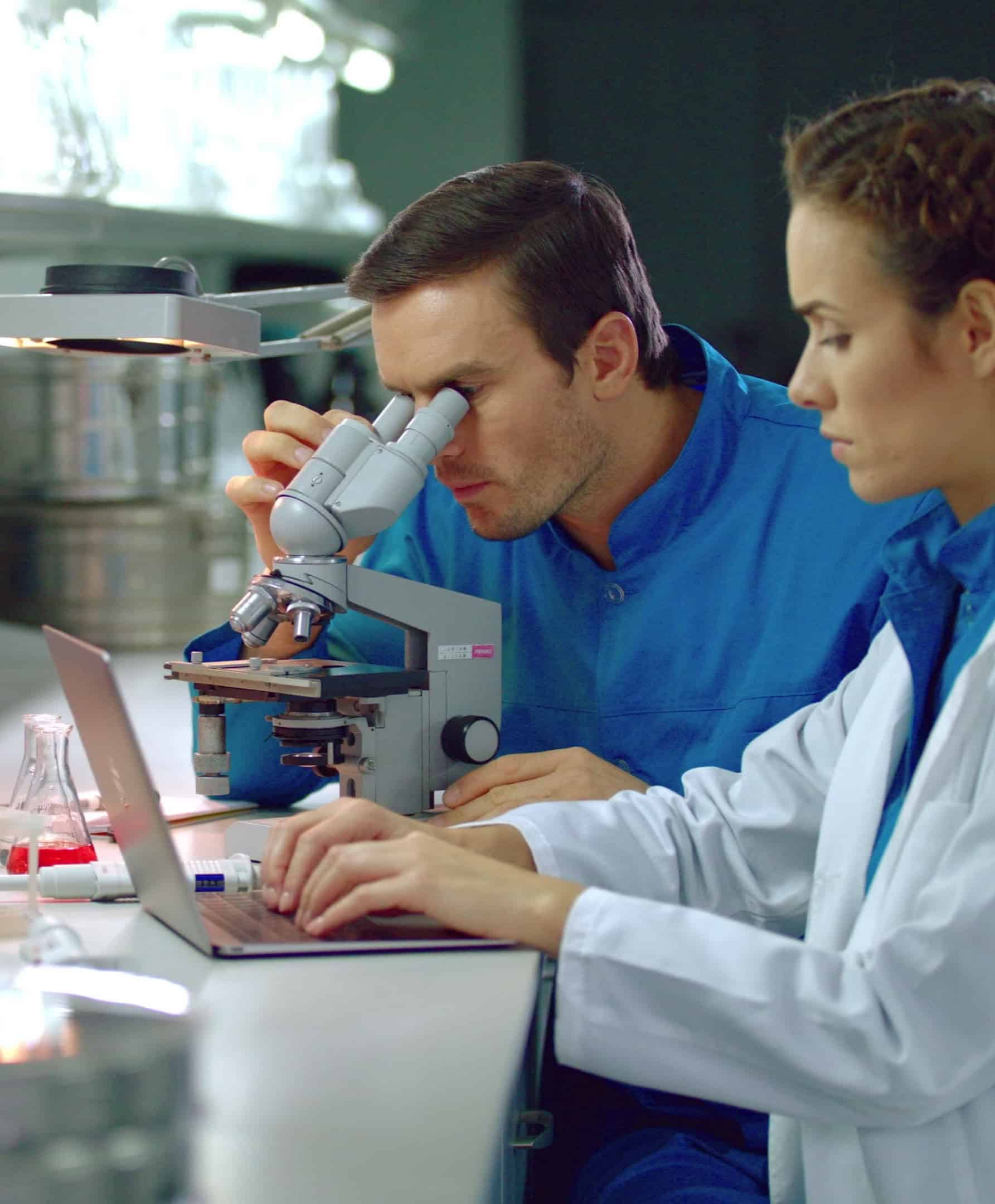 Twee onderzoekers werken achter een microscoop en met een laptop in een onderzoekslaboratorium van een CRO en CDMO.