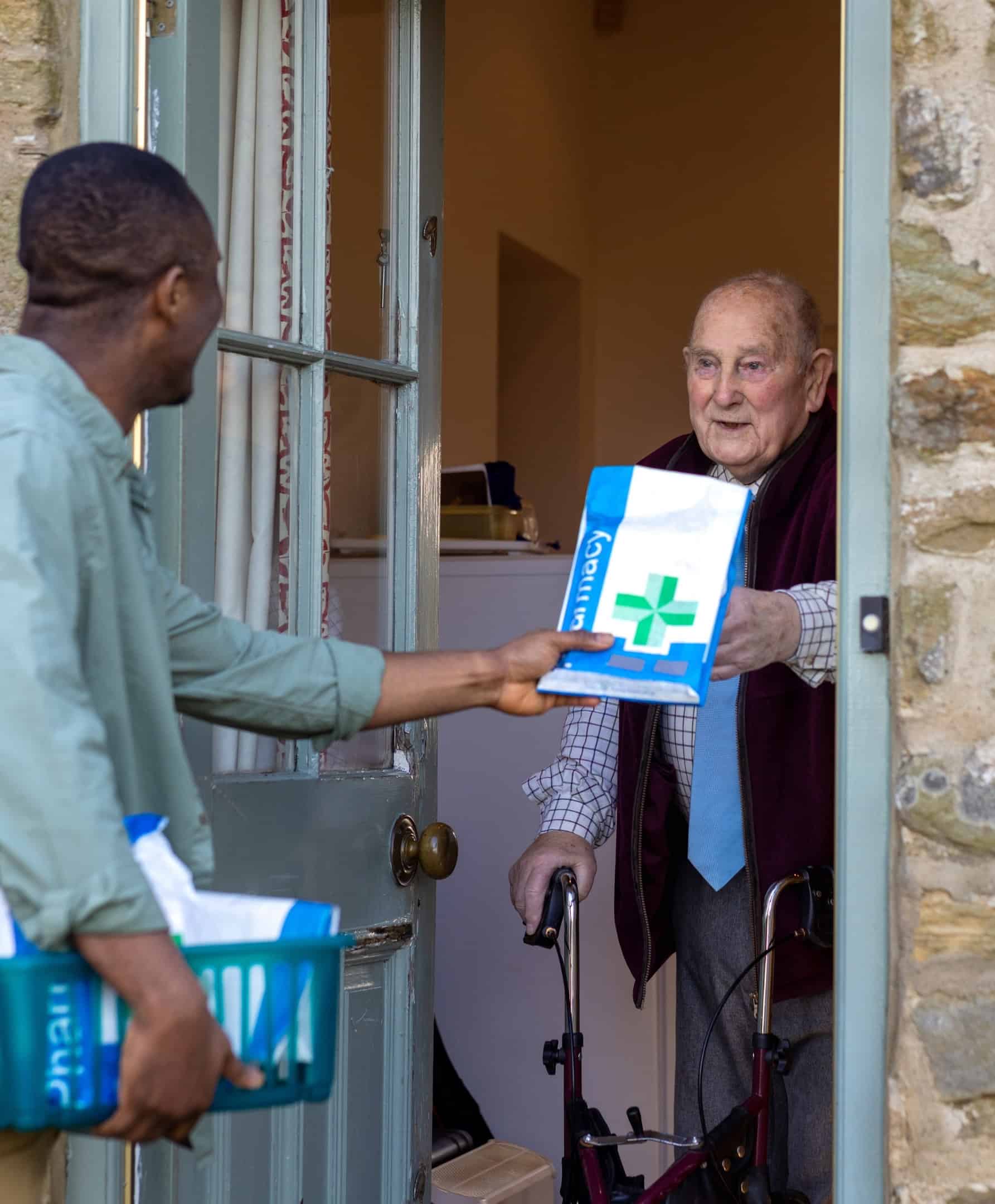 Een medewerker van een farmaceutisch distributiecentrum overhandigt een medicijnpakket aan een oudere patiënt bij diens voordeur.