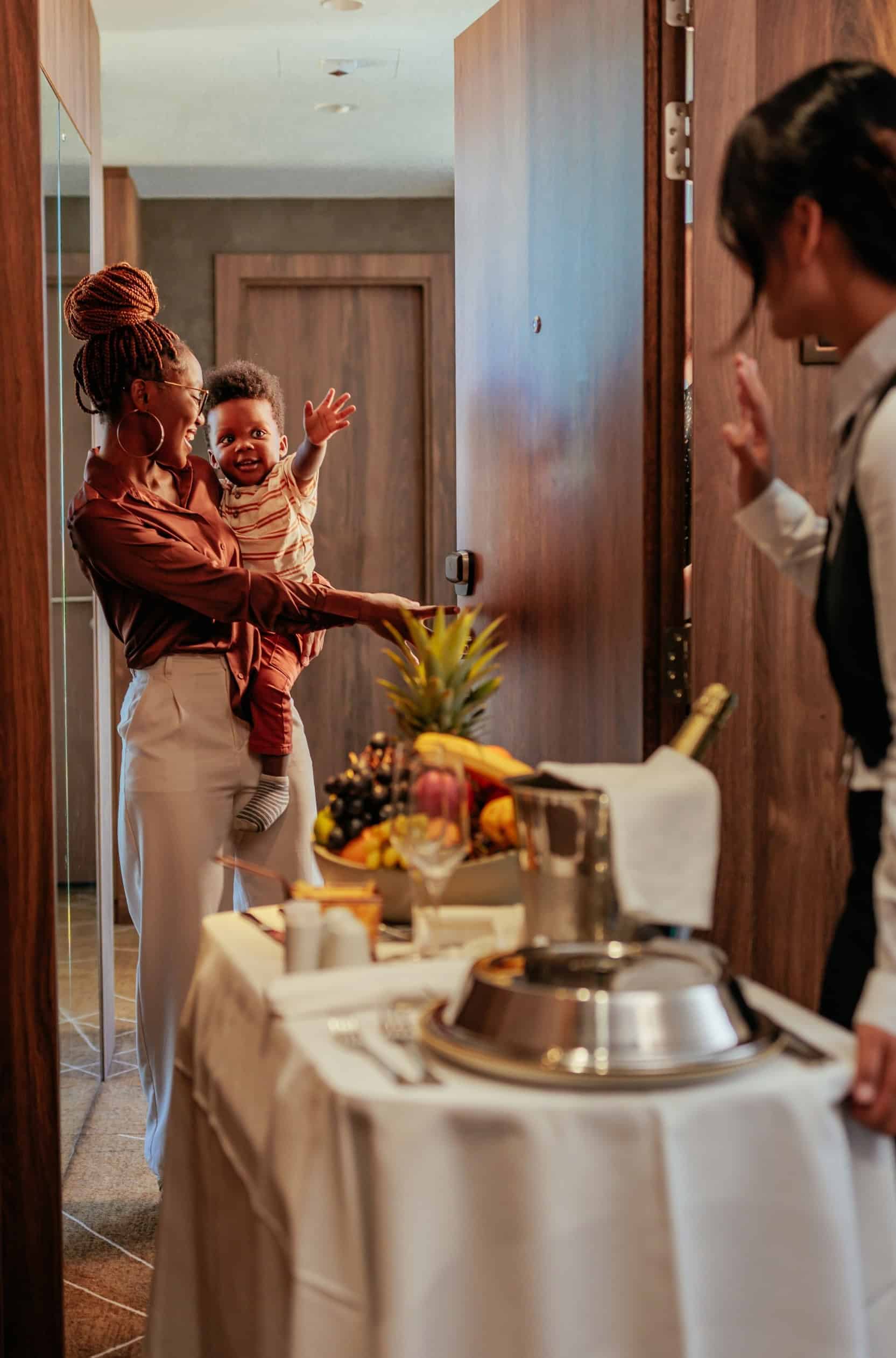 Hotel room service attendant delivering a food cart to a guest and her baby, representative of the consumer experience in the hospitality industry.