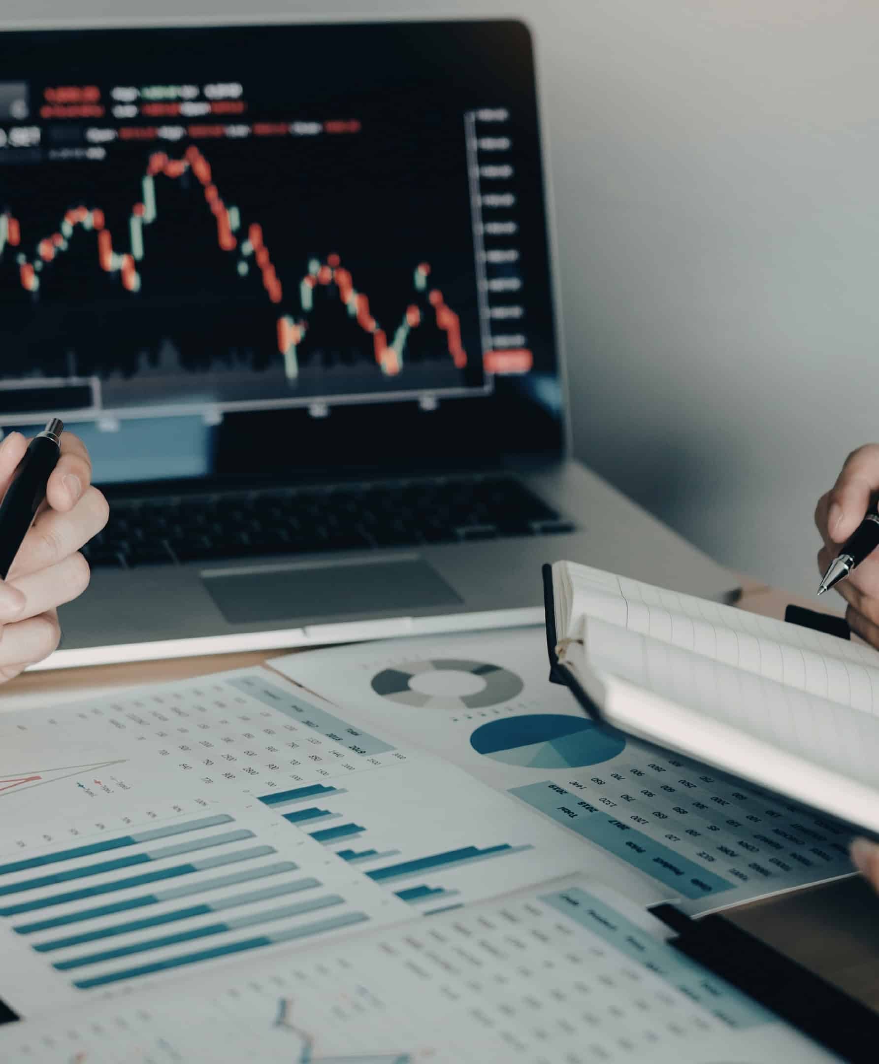 Analistas revisando datos financieros e informes analíticos junto con una pantalla de operaciones bursátiles.