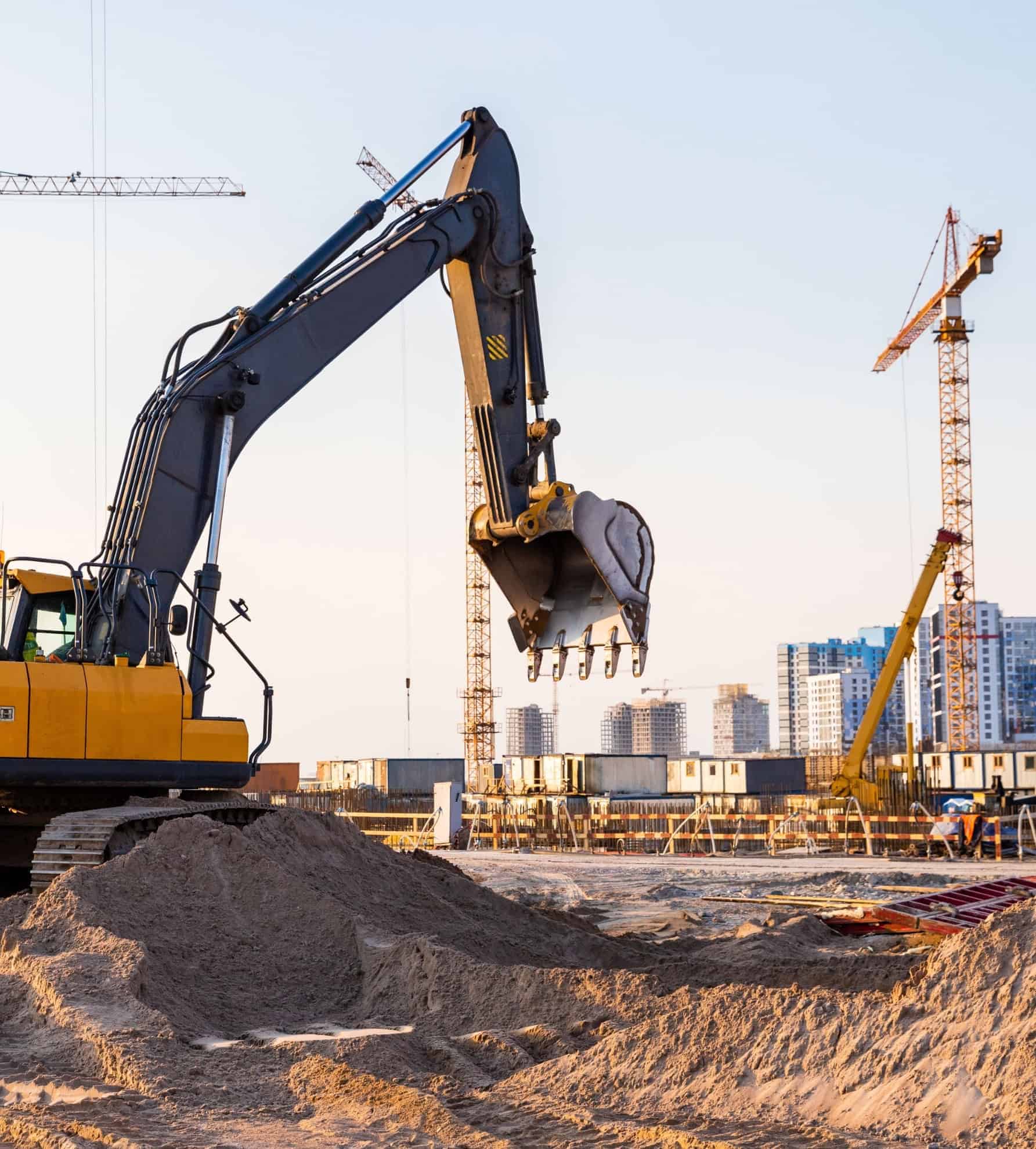 Een graafmachine aan het werk op een grote bouwplaats met torenkranen, wat een grootschalig project in de infrastructuur- en bouwsector illustreert.