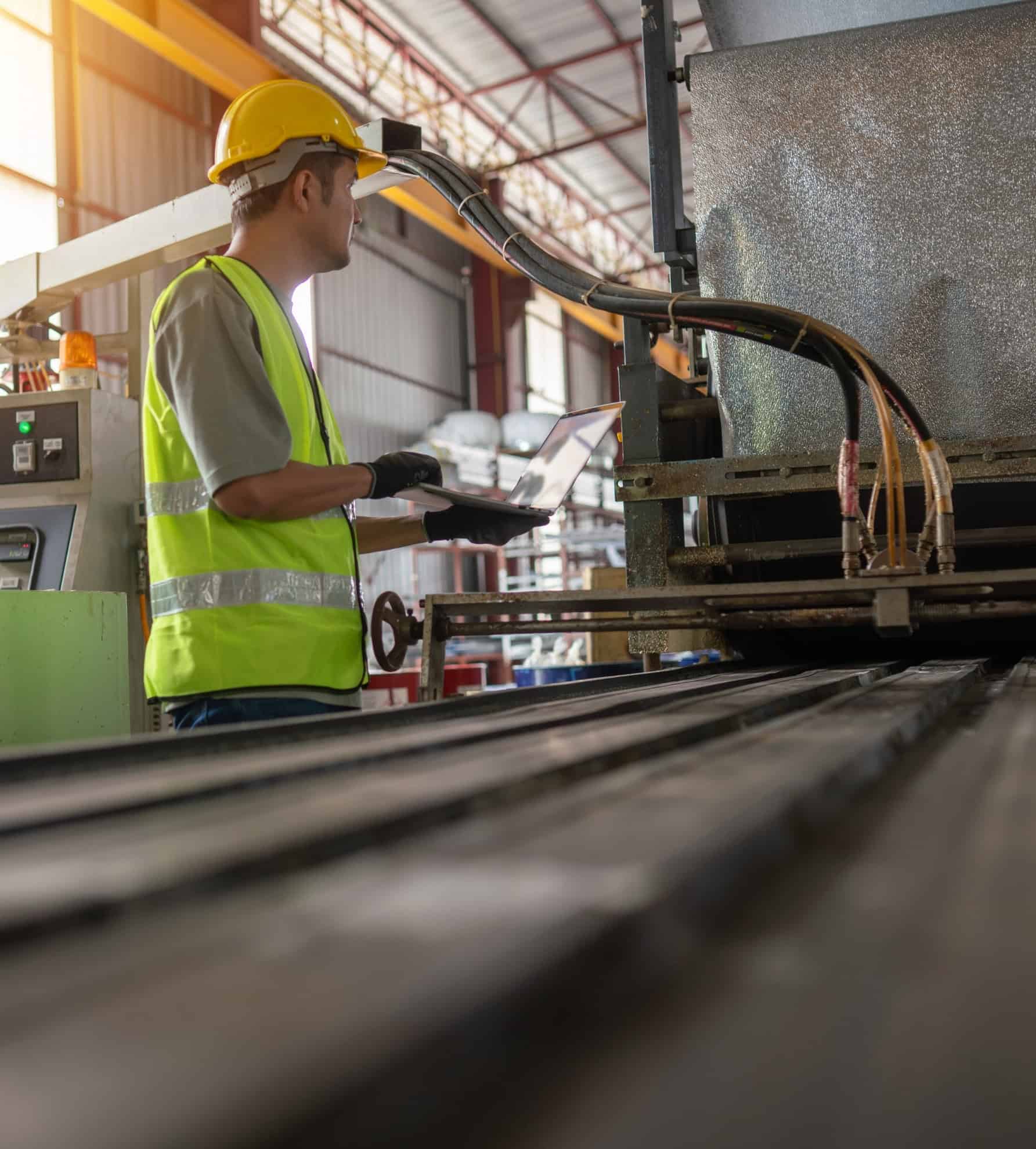 Een werknemer in een veiligheidsvest bedient industriële machines met een laptop en demonstreert daarmee het leiderschap van Industrie 4.0 in de productie.