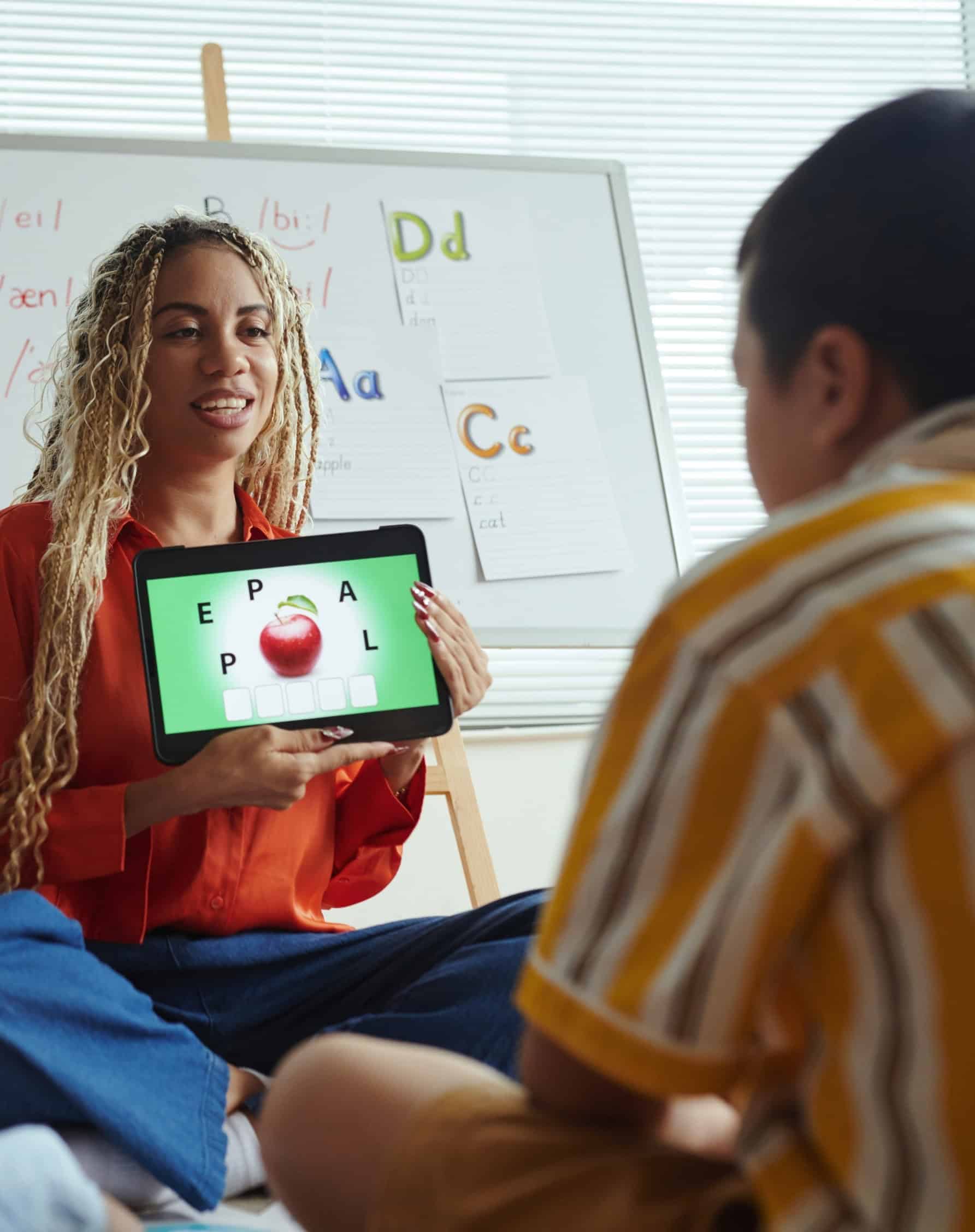 Teacher showing a young student an educational app on a tablet in a classroom.