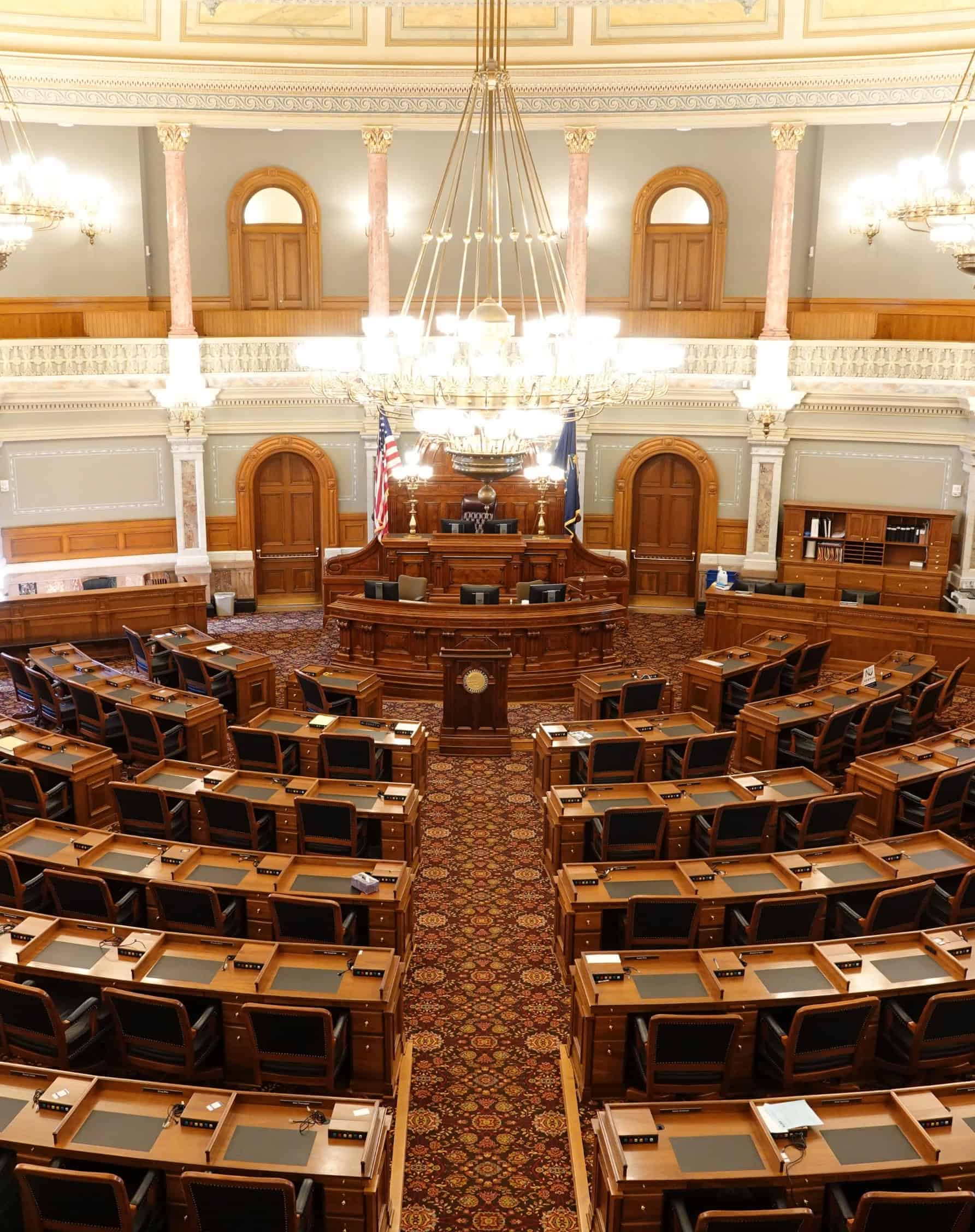 A grand legislative chamber with ornate wooden desks and an American flag representing government leadership.