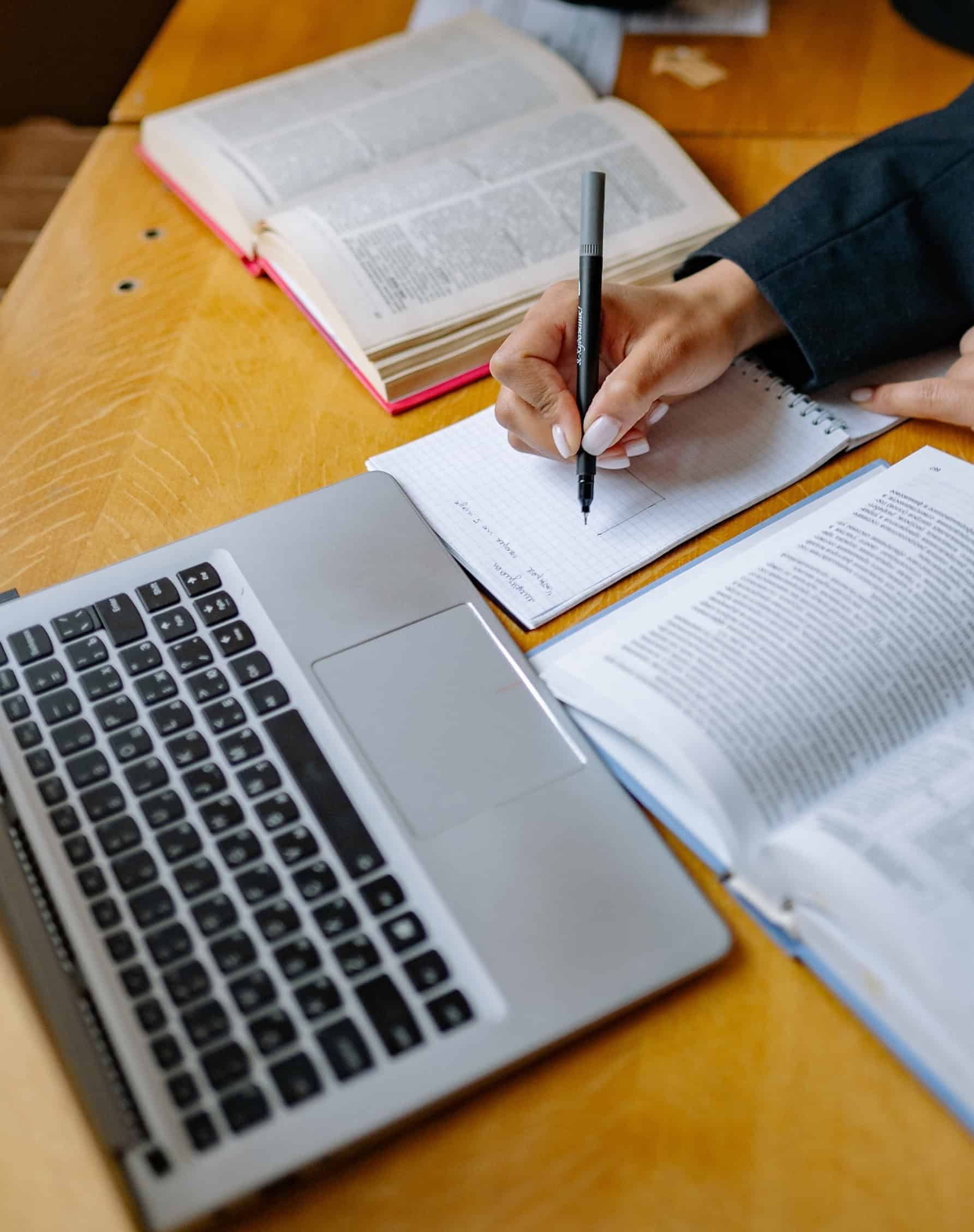 Scientific researcher writing notes for a scientific journal publication, with reference books and a laptop.