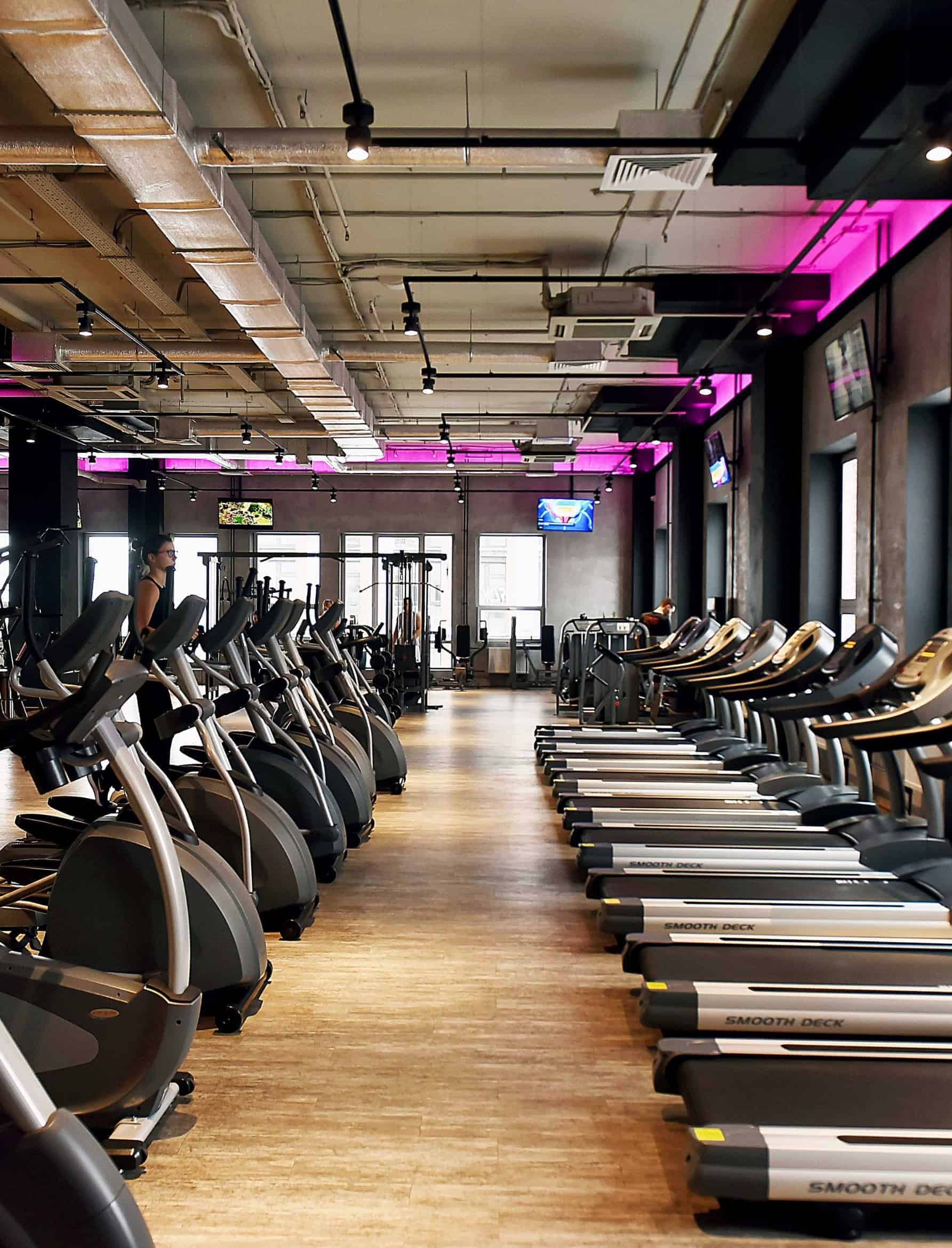 Modern gym environment with rows of treadmills and elliptical machines under purple mood lighting.